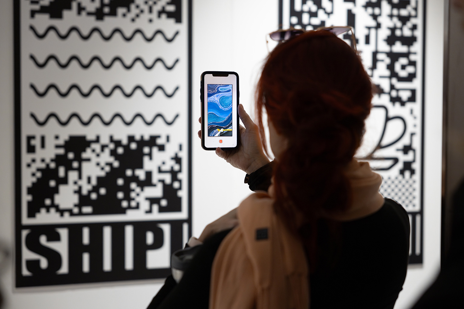 Sophie Kurtness - Festival Art Souterrain; Photo : Michael Patten Une femme tient sont smartphone devant une œuvre de l'artiste Sophie Kurtness. Un tableau différent de ce qu'il y a sur le mur apparaît sur l'écran